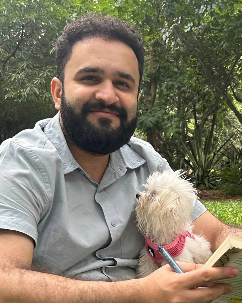 Akshat Kapur smiling, sitting on the grass with a small white dog and a book